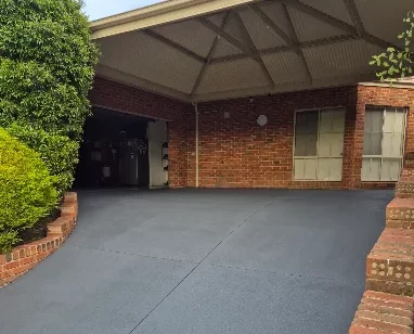 Wide grey spray-on concrete driveway with a smooth textured finish, curving toward a brick carport flanked by brick garden edging and lush greenery. Spray-on concrete driveway resurfacing by Spraycrete.